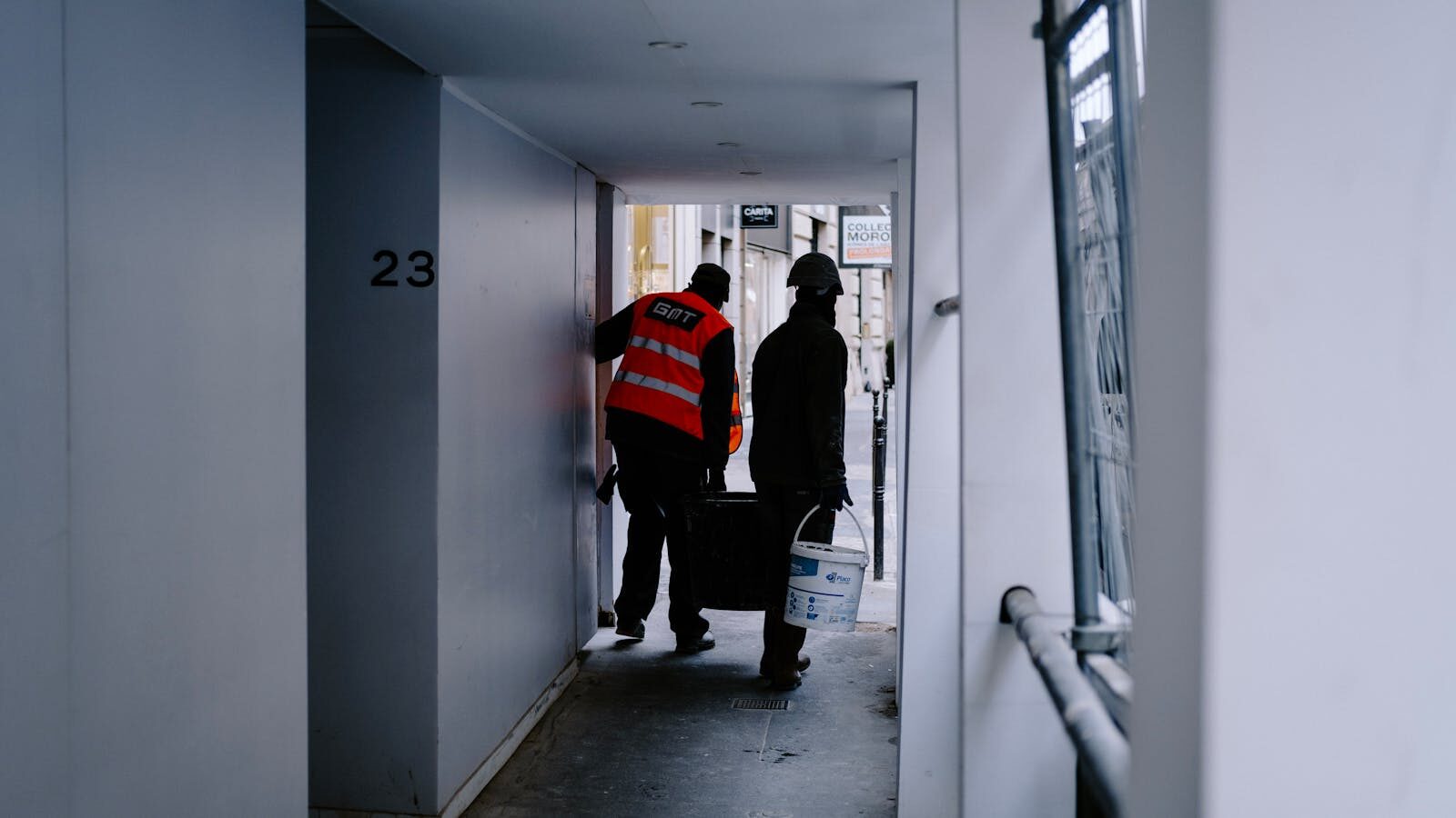 Two construction workers carrying materials in a city hallway.